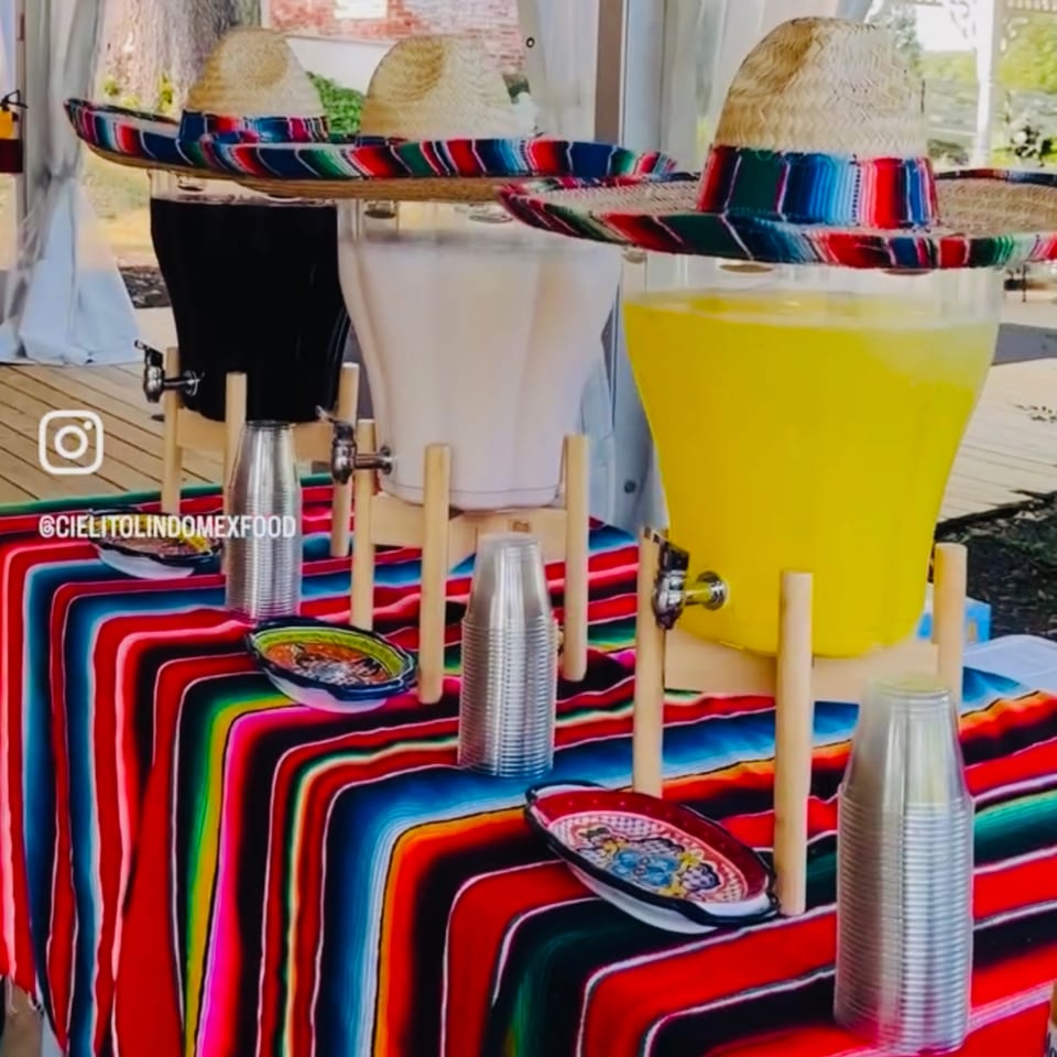Three beverage dispensers with sombreros, plastic cups, and decorative bowls on a colorful striped Mexican blanket.