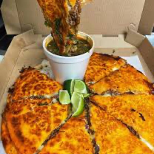 A birria pizza with a slice being dipped into a cup of consommé, garnished with lime wedges.