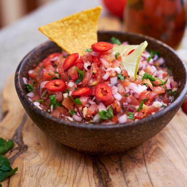 Fresh salsa in a dark bowl with a tortilla chip, lime, and chili slices on a wooden board.