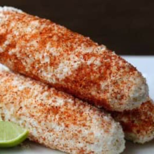 Two Mexican elotes, or grilled corn on the cob, covered in cheese and chili powder with a lime wedge.