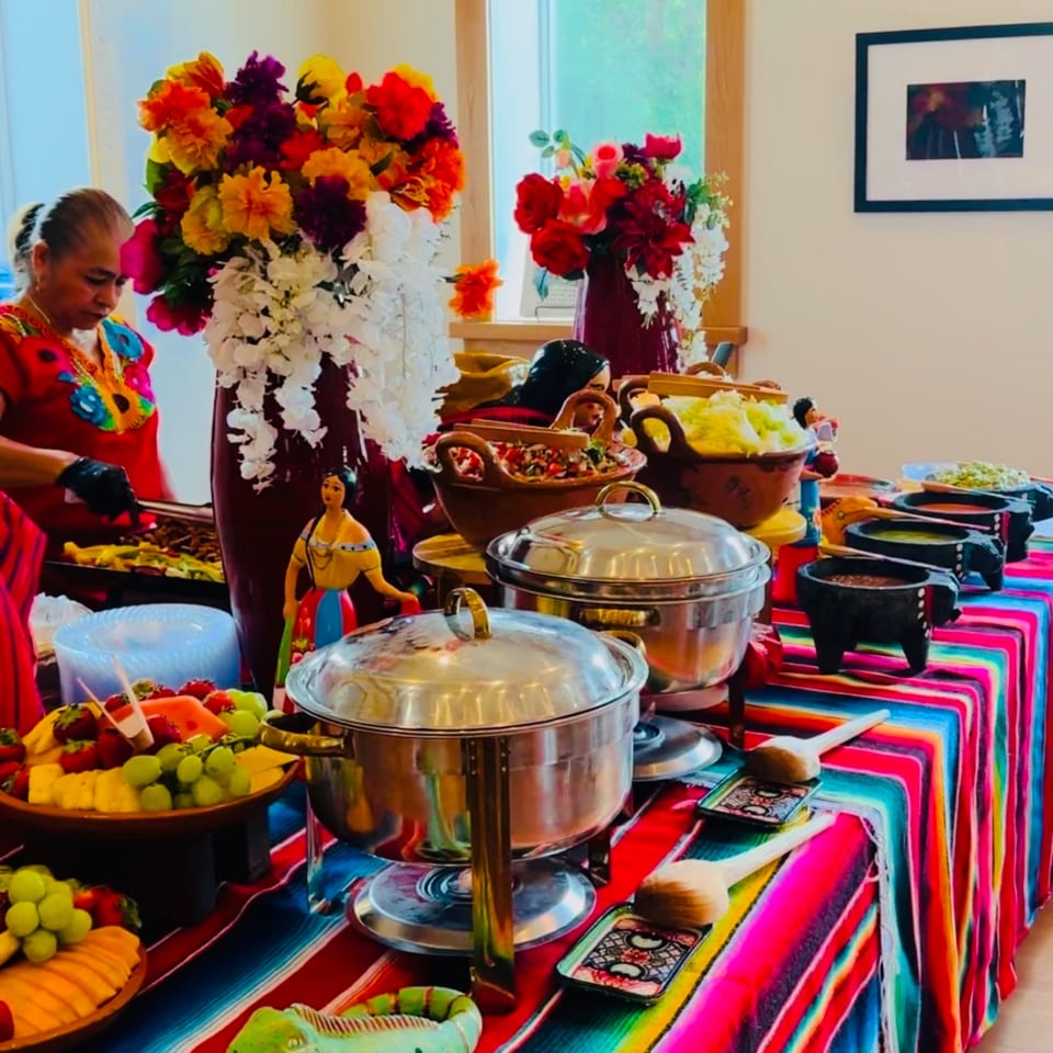 A vibrant Mexican buffet with a woman serving food, colorful flowers, and various dishes on a striped tablecloth.