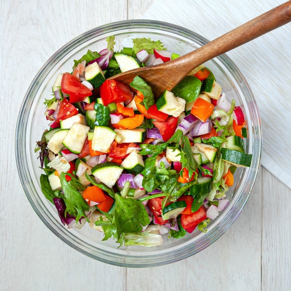 A vibrant Mexican salad in a glass bowl with a wooden spoon, featuring fresh mixed greens, tomatoes, zucchini, and red onion.