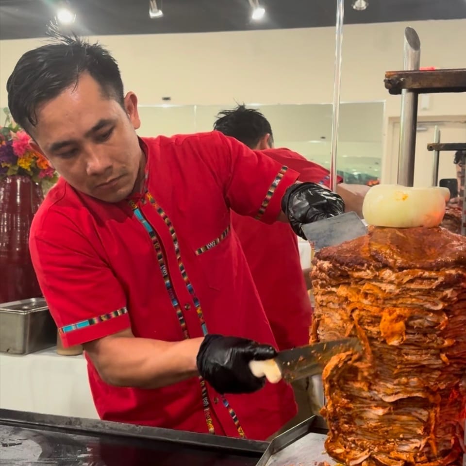A chef in a red shirt and black gloves carves al pastor meat from a vertical spit.