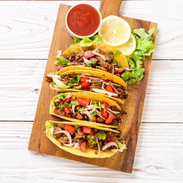 Four beef tacos with lettuce, tomatoes, and onions on a wooden board with salsa and lemon.
