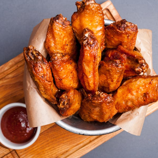 Crispy, golden-brown chicken wings in a metal bowl lined with parchment paper, with a side of red sauce.